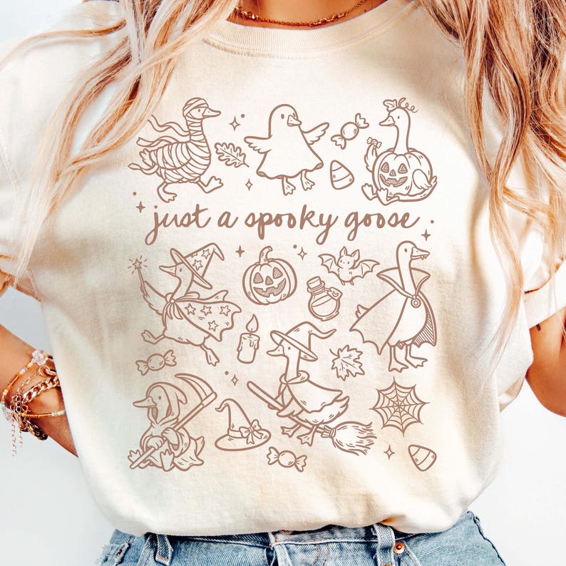 May include: Cream-colored sweatshirt featuring a brown graphic of Halloween-themed geese. The design includes geese dressed as mummies, witches, and vampires, along with pumpkins, candy, and the text "just a spooky goose."