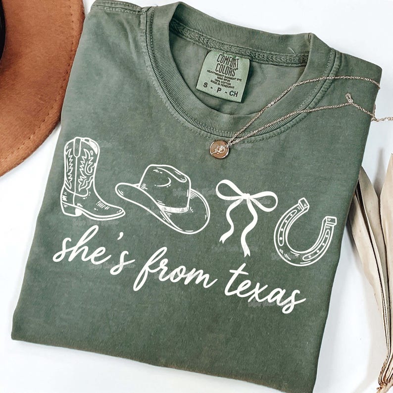 以下が含まれることがあります： 「she's from texas」の文字と、カウボーイブーツ、帽子、リボン、蹄鉄の白いイラストが描かれたオリーブグリーンのTシャツ。ペンダント付きのシルバーネックレスも写っています。