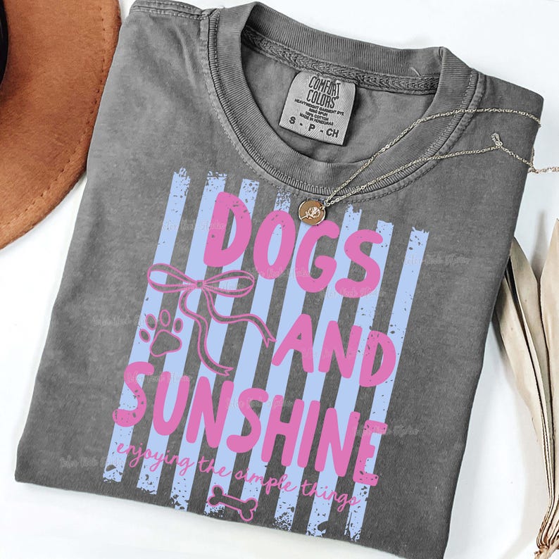 May include: Dark gray t-shirt with the text "DOGS AND SUNSHINE" in pink, set against blue vertical stripes. The shirt includes a paw print, a bow, and a bone graphic. A silver necklace with a pendant is visible.
