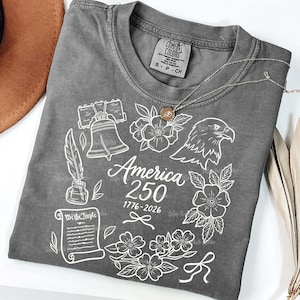May include: A gray Comfort Colors t-shirt with a white graphic design. The design includes the Liberty Bell, an eagle, flowers, a quill and ink, and the text "America 250 1776-2026". A silver necklace with a pendant is on the shirt.