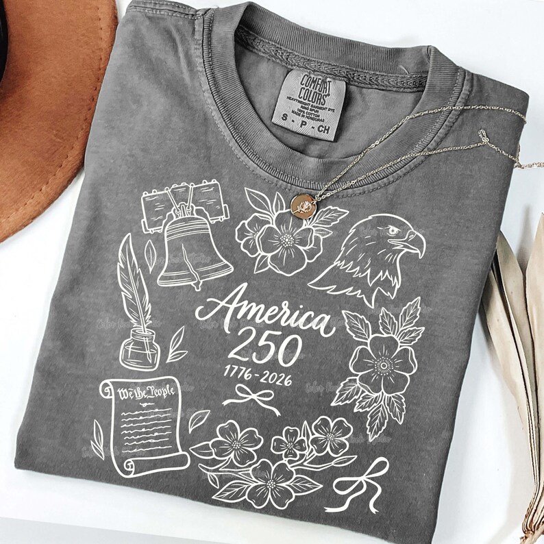 May include: A gray Comfort Colors t-shirt with a white graphic design. The design includes the Liberty Bell, an eagle, flowers, a quill and ink, and the text "America 250 1776-2026". A silver necklace with a pendant is on the shirt.