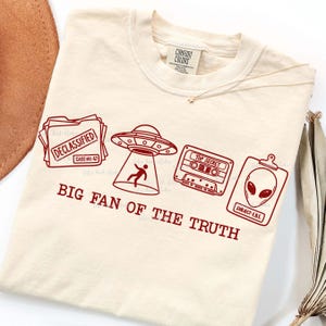 Könnte beinhalten: Cremefarbenes T-Shirt mit rotem Grafikdesign. Das Design zeigt eine Akte mit der Aufschrift "Declassified", ein UFO, eine Kassette mit der Aufschrift "Top Secret" und eine Alien-Akte. Der Text "BIG FAN OF THE TRUTH" ist darunter gedruckt.