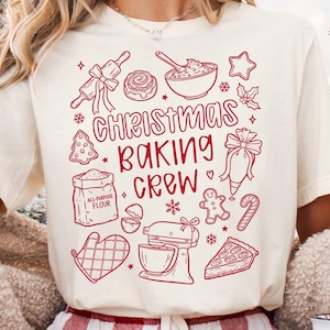 May include: Cream-colored t-shirt with a red graphic of baking-related illustrations and text. The design includes a rolling pin, bowl, gingerbread man, and the words "Christmas Baking Crew."