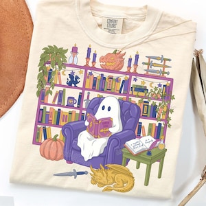 Peut inclure: Sweat-shirt crème avec une illustration fantaisiste. Un fantôme lit un livre dans un fauteuil violet devant une bibliothèque avec des citrouilles, des bougies, des épées et un dragon endormi. Le livre indique "Storm & Scale".