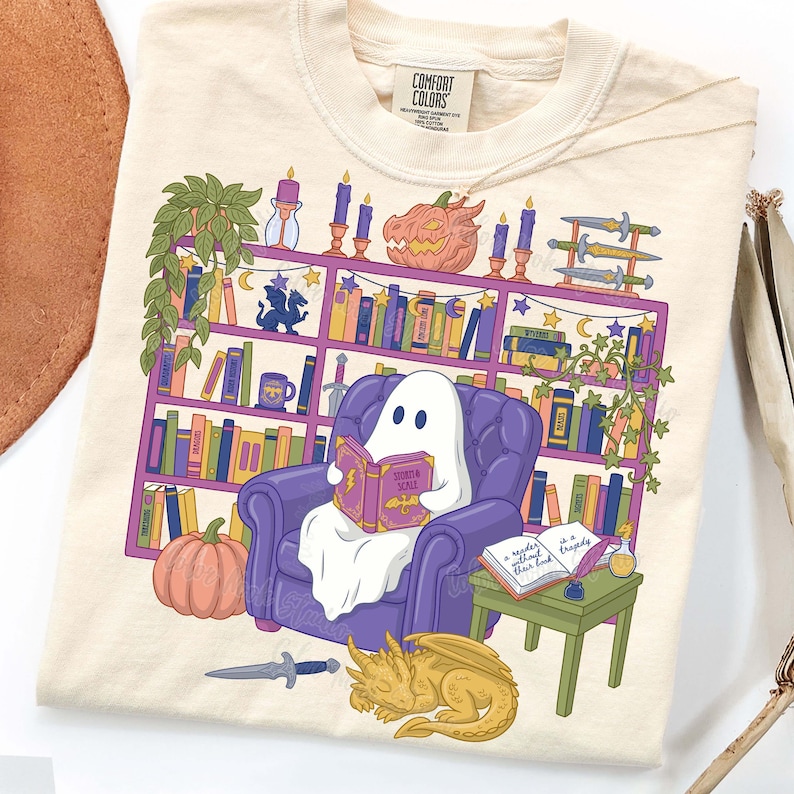 May include: Cream-coloured sweatshirt with a fantasy-themed illustration. A ghost reads a book in a purple armchair, in front of a bookshelf with pumpkins, candles, swords, and a sleeping dragon. The book reads "Storm & Scale".