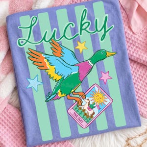 May include: A light purple t-shirt with a colorful graphic of a duck holding a tarot card that reads "The Sun". The word "Lucky" is written in green script above the duck. The shirt has green vertical stripes.