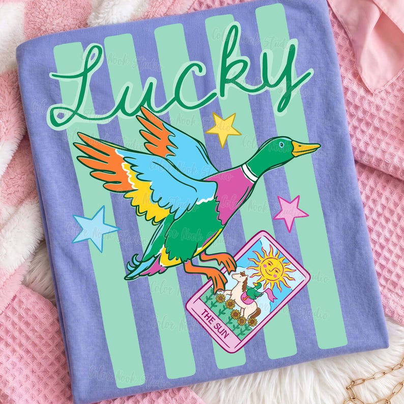 May include: A light purple t-shirt with a colorful graphic of a duck holding a tarot card that reads "The Sun". The word "Lucky" is written in green script above the duck. The shirt has green vertical stripes.