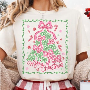 Pode incluir: Camiseta creme com um desenho de árvore de Natal feito de laços rosa e verdes. As palavras "Merry Christmas" estão escritas em rosa abaixo da árvore. O desenho é emoldurado por uma borda verde com bengalas doces e flocos de neve.