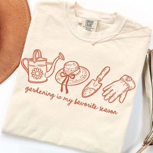 May include: Cream-colored t-shirt with a brown graphic of gardening tools: a watering can, hat, trowel, and gloves. The text "gardening is my favorite season" is printed below the illustrations. The shirt is made of a soft material.
