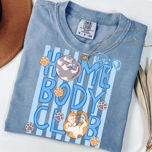May include: A blue Comfort Colors t-shirt with the words "HOME BODY CLUB" in light blue, featuring illustrations of two cats and paw prints. A gold necklace with a pendant and a brown hat are also visible.