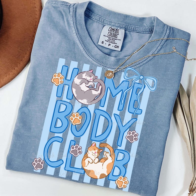 May include: A blue Comfort Colors t-shirt with the words "HOME BODY CLUB" in light blue, featuring illustrations of two cats and paw prints. A gold necklace with a pendant and a brown hat are also visible.
