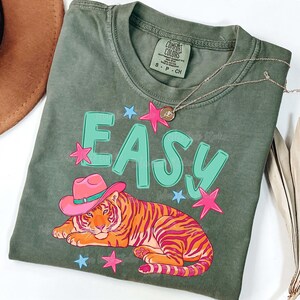 Puede incluir: Una camiseta verde salvia con la palabra "EASY" en letras turquesas grandes. Un tigre de dibujos animados con un sombrero de vaquero rosa est&aacute; debajo de la palabra. Estrellas rosas y azules rodean el texto y el tigre. Un collar plateado est&aacute; sobre la camisa.