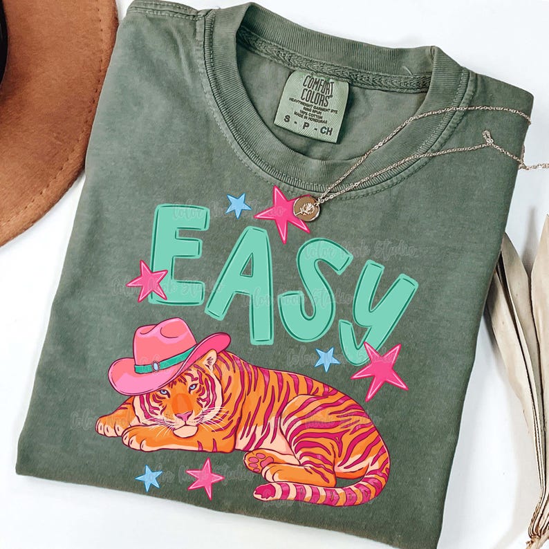 Puede incluir: Una camiseta verde salvia con la palabra "EASY" en letras turquesas grandes. Un tigre de dibujos animados con un sombrero de vaquero rosa est&aacute; debajo de la palabra. Estrellas rosas y azules rodean el texto y el tigre. Un collar plateado est&aacute; sobre la camisa.