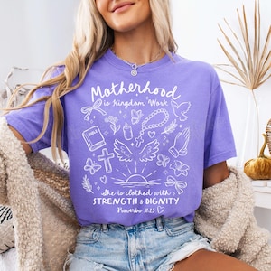 May include: Purple t-shirt with a white graphic design that says "Motherhood is Kingdom Work." The graphic also includes a Bible, a cross, a dove, a crown of thorns, a candle, and a heart. The text at the bottom says "She is clothed with STRENGTH & DIGNITY Proverbs 31:25."