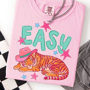 Puede incluir: Camiseta rosa claro con la palabra "EASY" en letras turquesas. Un tigre con un sombrero de vaquero rosa est&aacute; debajo de la palabra. Estrellas rosas y azules rodean el texto y el tigre. Una cadena de plata cuelga sobre la camiseta.