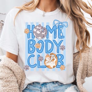 May include: White t-shirt with a blue and white striped design and the words "HOME BODY CLUB" in blue. The shirt features illustrations of cats and paw prints.