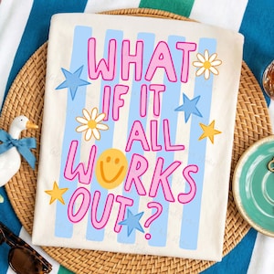 May include: Off-white t-shirt with the text "WHAT IF IT ALL WORKS OUT?" in pink outlined letters. The design includes blue stripes, stars, daisies, and a yellow smiley face. The shirt is on a woven mat.