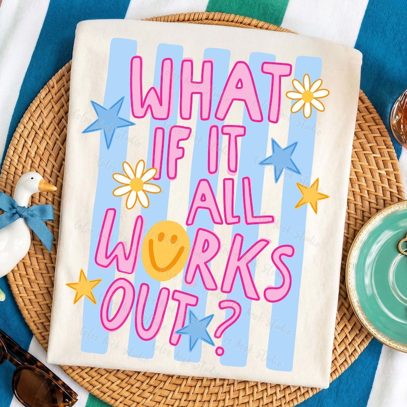May include: Off-white t-shirt with the text "WHAT IF IT ALL WORKS OUT?" in pink outlined letters. The design includes blue stripes, stars, daisies, and a yellow smiley face. The shirt is on a woven mat.