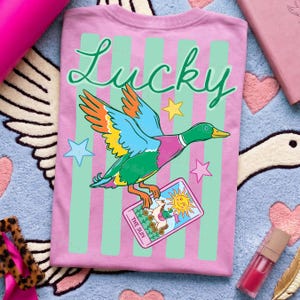 May include: Pink t-shirt with a green and white striped design. The shirt features the word "Lucky" in green script and a colorful illustration of a duck holding a tarot card that says "The Sun".