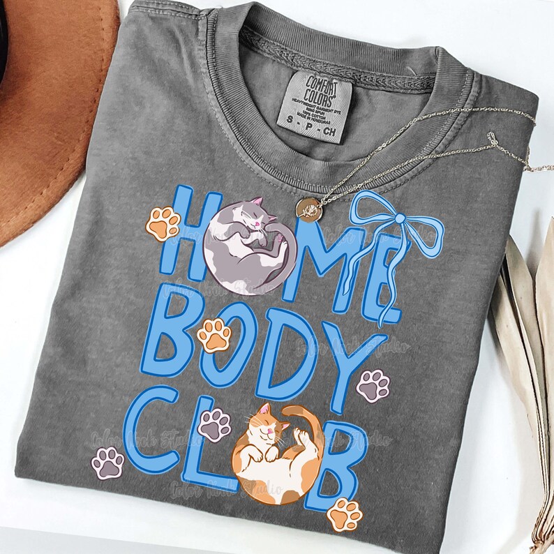 May include: A gray Comfort Colors t-shirt with the words "HOME BODY CLUB" in blue, featuring cat illustrations and paw prints. A silver necklace and a brown hat are also visible.