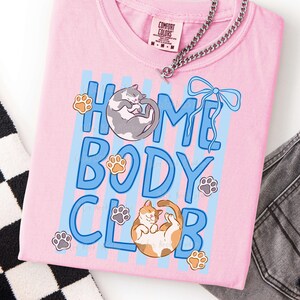 May include: A light pink t-shirt with the words "HOME BODY CLUB" in blue, featuring illustrations of cats and paw prints. A silver chain necklace and a Comfort Colors tag are visible. The shirt is folded.