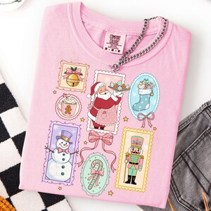 May include: A light pink t-shirt featuring a festive Christmas design. The shirt showcases various holiday-themed illustrations, including Santa Claus, a snowman, a nutcracker, and a stocking filled with gifts. The shirt has a Comfort Colors tag.