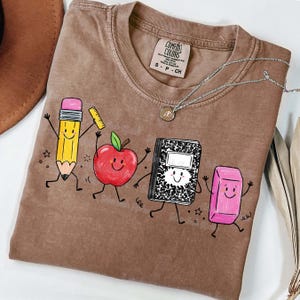 May include: Brown Comfort Colors t-shirt featuring a playful graphic of a pencil, apple, notebook, and eraser with smiling faces. The shirt has a crew neck and short sleeves. A silver necklace with a pendant is also visible.