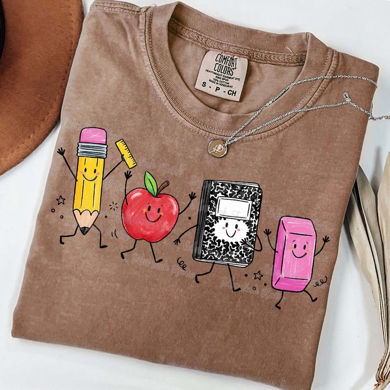 May include: Brown Comfort Colors t-shirt featuring a playful graphic of a pencil, apple, notebook, and eraser with smiling faces. The shirt has a crew neck and short sleeves. A silver necklace with a pendant is also visible.