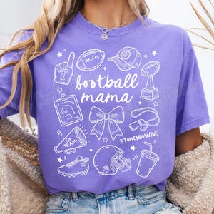 May include: Purple t-shirt with a white graphic design that says "football mama" and includes various football-related icons, including a football, a whistle, a trophy, a cheerleader's bow, and a football helmet.