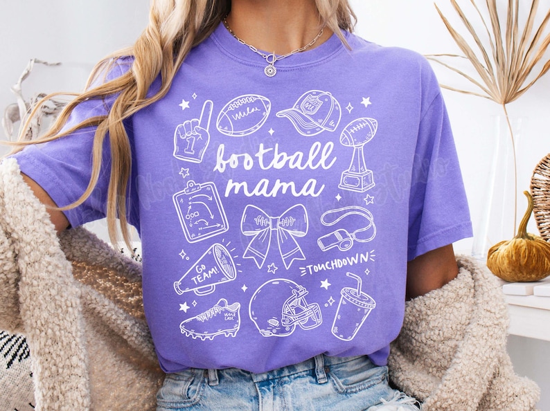 May include: Purple t-shirt with a white graphic design that says "football mama" and includes various football-related icons, including a football, a whistle, a trophy, a cheerleader's bow, and a football helmet.