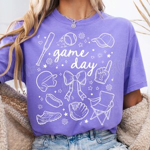 K&ouml;nnte beinhalten: Lilafarbenes T-Shirt mit einem wei&szlig;en Grafikdesign von Baseball-Motiven, darunter ein Schl&auml;ger, ein Ball, ein Handschuh, eine M&uuml;tze und der Text "game day".