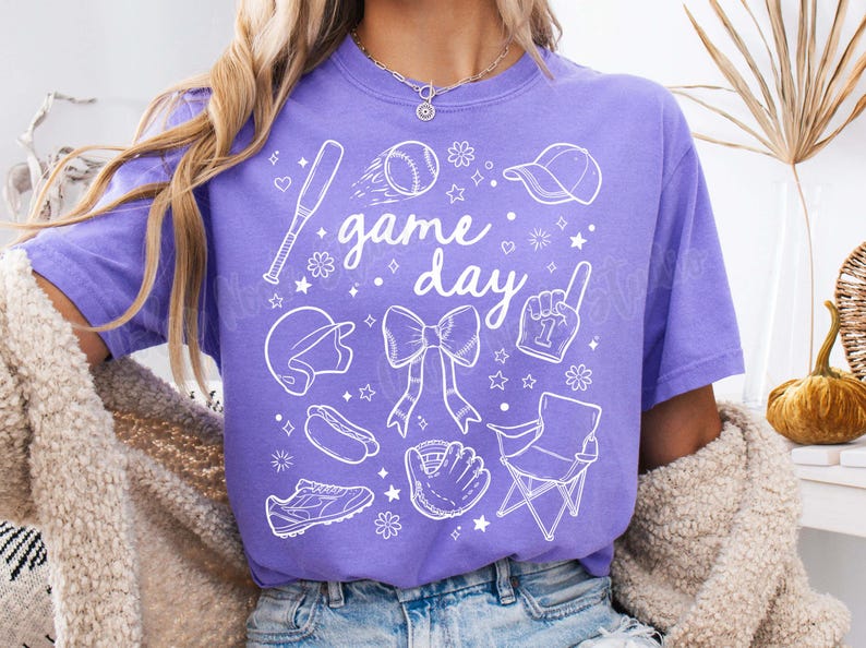 K&ouml;nnte beinhalten: Lilafarbenes T-Shirt mit einem wei&szlig;en Grafikdesign von Baseball-Motiven, darunter ein Schl&auml;ger, ein Ball, ein Handschuh, eine M&uuml;tze und der Text "game day".