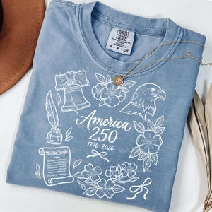 May include: A blue Comfort Colors t-shirt with a white graphic design. The design includes the Liberty Bell, an eagle, flowers, and the text "America 250 1776-2026". A gold necklace is draped across the shirt.