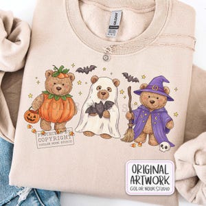 Op de afbeelding: Een beige sweater met ronde hals voorzien van een afbeelding van drie teddyberen in Halloween-kostuums: &eacute;&eacute;n als pompoen, &eacute;&eacute;n als spook met een vleermuis en &eacute;&eacute;n als tovenaar met een bezem, hoed en mantel, omringd door kleine sterren.