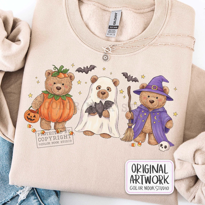Op de afbeelding: Een beige sweater met ronde hals voorzien van een afbeelding van drie teddyberen in Halloween-kostuums: &eacute;&eacute;n als pompoen, &eacute;&eacute;n als spook met een vleermuis en &eacute;&eacute;n als tovenaar met een bezem, hoed en mantel, omringd door kleine sterren.