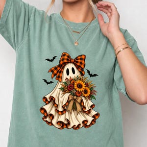 Peut inclure: Un t-shirt à manches courtes vert sauge avec un motif représentant un fantôme blanc portant un nœud à carreaux orange et noir, tenant un bouquet de tournesols et de fleurs d'automne, avec trois chauves-souris noires en arrière-plan.