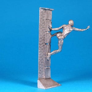 K&ouml;nnte beinhalten: Eine silberne Figur eines Menschen, der eine Ziegelmauer hochklettert. Die Person streckt einen Arm aus und hat ein Bein hinter sich gebeugt. Die Figur steht auf einem kleinen Sockel.