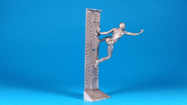 K&ouml;nnte beinhalten: Eine silberne Figur eines Menschen, der eine Ziegelmauer hochklettert. Die Person streckt einen Arm aus und hat ein Bein hinter sich gebeugt. Die Figur steht auf einem kleinen Sockel.