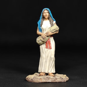 May include: A figurine of a woman in a white dress and blue head covering, holding a bundle of wheat. The figure has a red sash and stands on a textured base. The background is black.