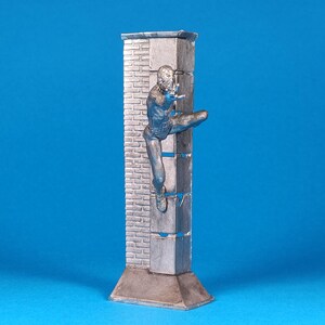 K&ouml;nnte beinhalten: Eine silberfarbene Figur eines Menschen, der eine Ziegelmauer erklimmt. Die Figur ist in einer dynamischen Aktionsstellung dargestellt, mit einem ausgestreckten Bein und dem anderen Bein, das im Knie gebeugt ist. Die Mauer besteht aus Ziegeln und hat eine raue Textur. Die Figur steht auf einem Sockel, der ebenfalls aus Silber ist.