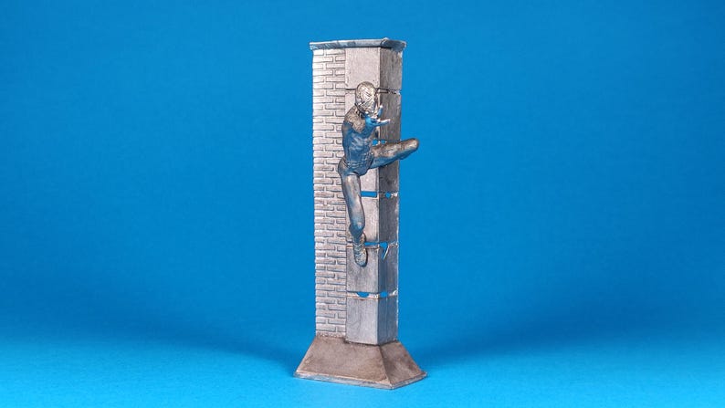 K&ouml;nnte beinhalten: Eine silberfarbene Figur eines Menschen, der eine Ziegelmauer erklimmt. Die Figur ist in einer dynamischen Aktionsstellung dargestellt, mit einem ausgestreckten Bein und dem anderen Bein, das im Knie gebeugt ist. Die Mauer besteht aus Ziegeln und hat eine raue Textur. Die Figur steht auf einem Sockel, der ebenfalls aus Silber ist.