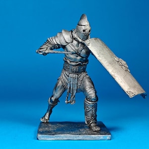 Puede incluir: Figura de gladiador plateada, posando con espada y escudo. La figura viste una armadura detallada, incluyendo casco, protectores de hombros y protección para las piernas. El escudo tiene elementos decorativos. Sobre un fondo azul.