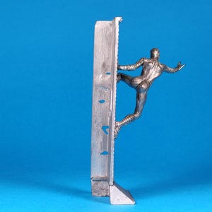 K&ouml;nnte beinhalten: Eine silberne Figur eines Menschen, der eine Wand hochklettert. Die Figur ist in einer dynamischen Pose dargestellt, mit einem ausgestreckten Bein und dem anderen Bein, das im Knie gebeugt ist. Die Wand besteht aus einem strukturierten Material und hat eine rechteckige Form.
