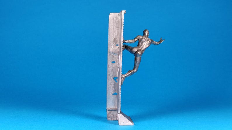 K&ouml;nnte beinhalten: Eine silberne Figur eines Menschen, der eine Wand hochklettert. Die Figur ist in einer dynamischen Pose dargestellt, mit einem ausgestreckten Bein und dem anderen Bein, das im Knie gebeugt ist. Die Wand besteht aus einem strukturierten Material und hat eine rechteckige Form.