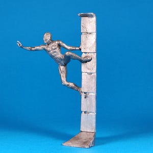 K&ouml;nnte beinhalten: Eine silberne Figur eines Menschen, der eine Wand klettert. Die Person befindet sich in der Luft, streckt eine Hand aus und h&auml;lt sich mit der anderen an der Wand fest. Die Wand besteht aus einer Reihe von rechteckigen Bl&ouml;cken.