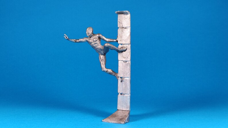 K&ouml;nnte beinhalten: Eine silberne Figur eines Menschen, der eine Wand klettert. Die Person befindet sich in der Luft, streckt eine Hand aus und h&auml;lt sich mit der anderen an der Wand fest. Die Wand besteht aus einer Reihe von rechteckigen Bl&ouml;cken.