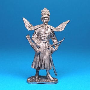May include: A silver-colored miniature figurine of a historical figure, possibly a soldier or nobleman, with elaborate attire. The figure has a feathered headdress, wing-like adornments, and holds a bow and quiver. The figurine is on a blue background.