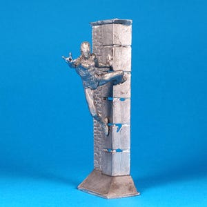 K&ouml;nnte beinhalten: Eine silberne Figur eines Menschen, der eine Wand klettert. Die Figur ist in einer dynamischen Kletterposition dargestellt, mit einem ausgestreckten Bein und dem anderen gebeugt. Die Wand besteht aus gestapelten Bl&ouml;cken, und die Figur klammert sich an die Seite der Wand.