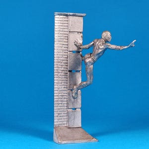 K&ouml;nnte beinhalten: Eine silberne Figur eines Menschen, der eine Ziegelmauer erklimmt. Die Person streckt die rechte Hand aus und das linke Bein ist im Knie gebeugt. Die Figur steht auf einem Sockel mit einem Ziegelmauerdesign.