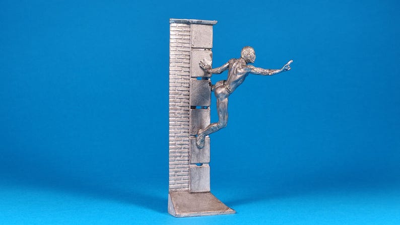 K&ouml;nnte beinhalten: Eine silberne Figur eines Menschen, der eine Ziegelmauer erklimmt. Die Person streckt die rechte Hand aus und das linke Bein ist im Knie gebeugt. Die Figur steht auf einem Sockel mit einem Ziegelmauerdesign.